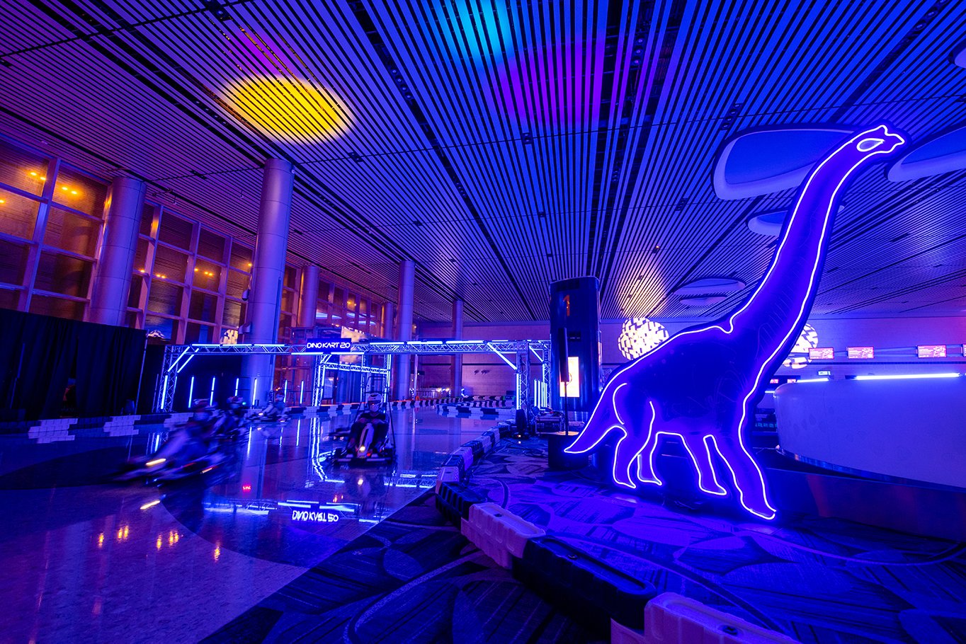 Racer speeding past check-in rows in Terminal 4 Departure Hall at Dino Kart – a dinosaur-themed indoor electric go-kart track with neon lights