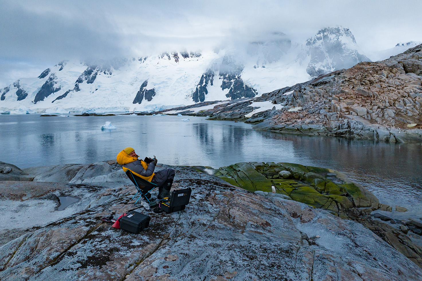 Bertie Gregory flies a drone sitting on a rock against the backdrop of a steep snowy mountain range.